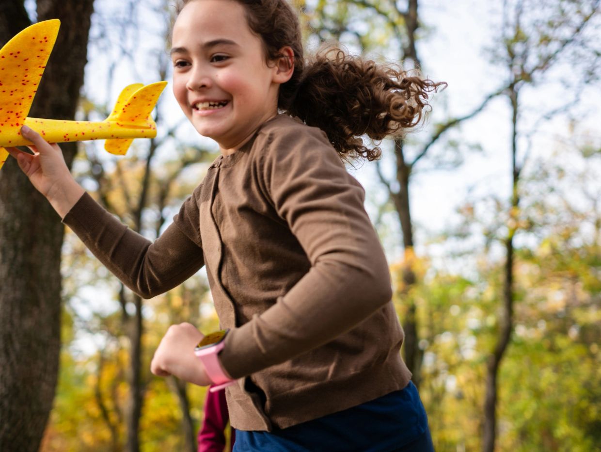 A happy girl running with a yellow toy airplane outdoors.