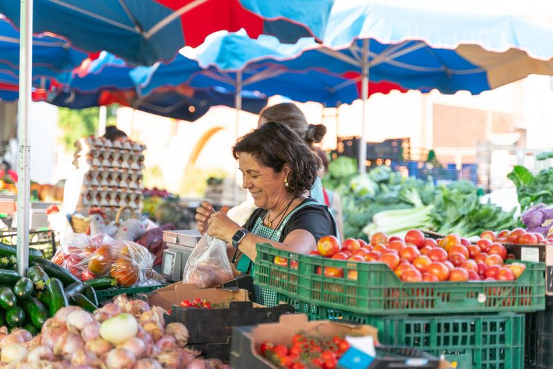 Smiling greengrocer weighing fresh produce at a farmers market stall, surrounded by colorful fruits and vegetables