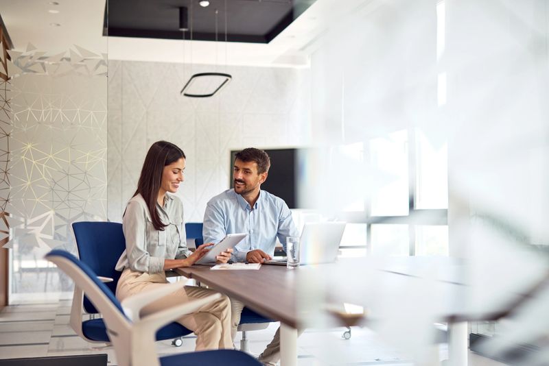 Two business partners are sitting at a table in a modern office, working together on a project using a digital tablet and laptop, discussing ideas and strategies