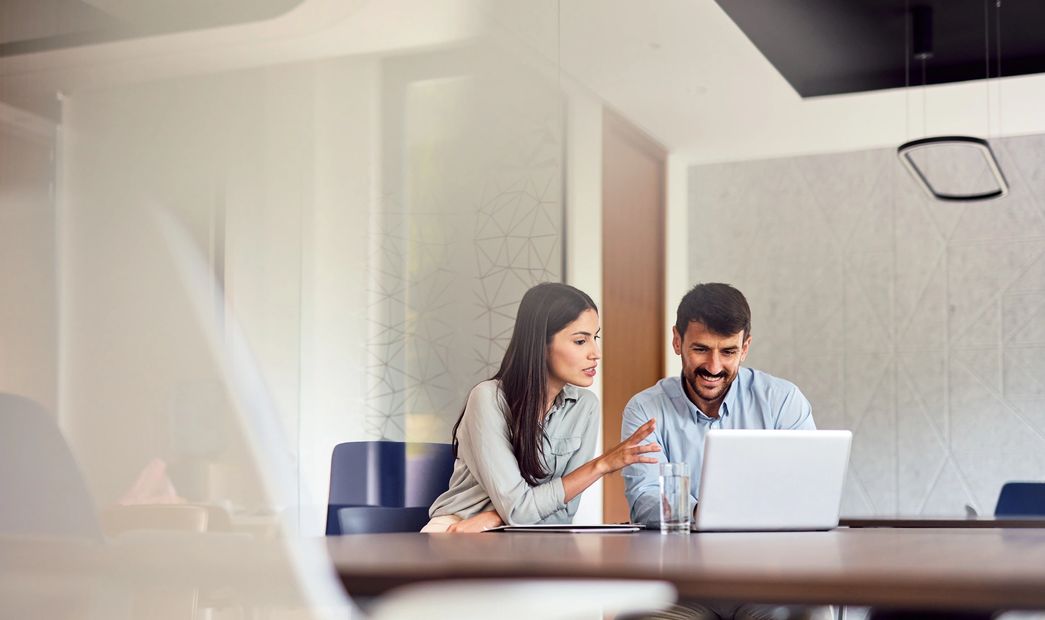 Two colleagues collaborating in a modern office, focused on a laptop screen.