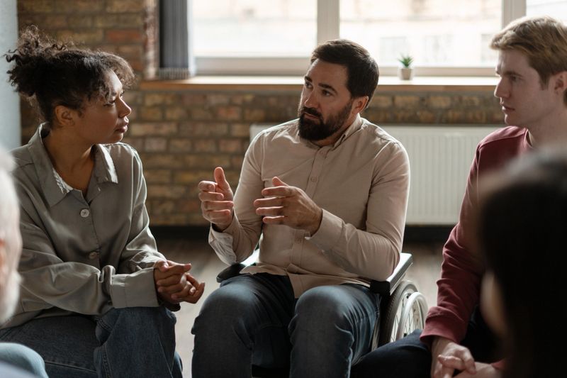 Man in a wheelchair gesturing and engaging in discussion with others during a supportive group therapy session, fostering connection and understanding