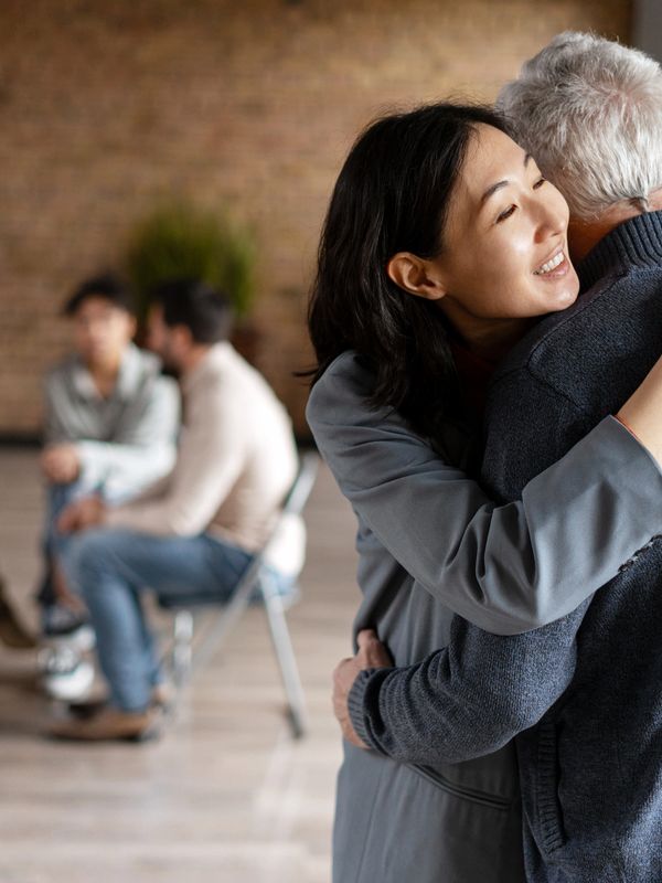 Two people warmly embracing in foreground with a conversation happening in the background.