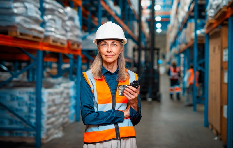 A confident warehouse worker, dressed in safety gear, supervises operations in a well-organized logistics storage facility. The busy environment signifies teamwork, efficiency, and the importance of safety in modern supply chain processes.