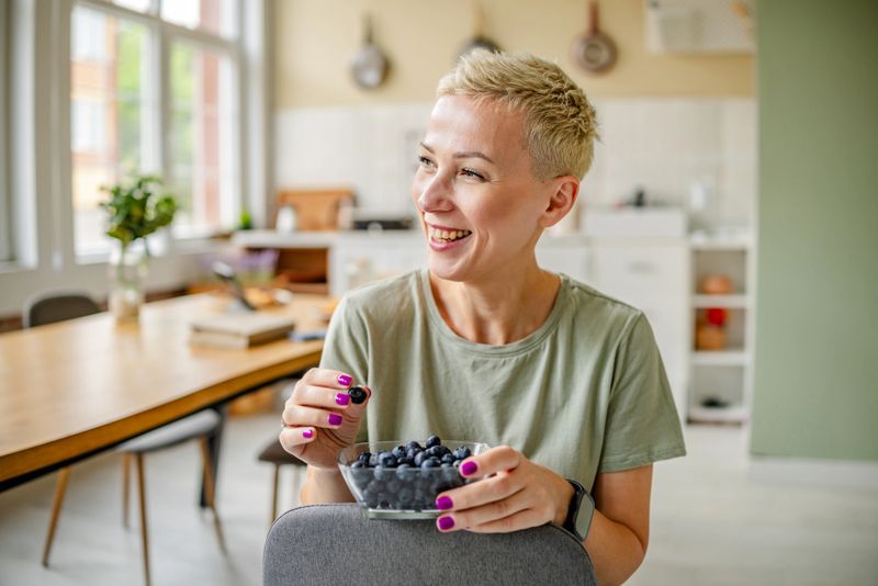 A smiling woman holds a bowl of fresh blueberries in a bright kitchen. She embodies a healthy lifestyle and enjoys snacking on nutritious fruits, celebrating the joy of active living.