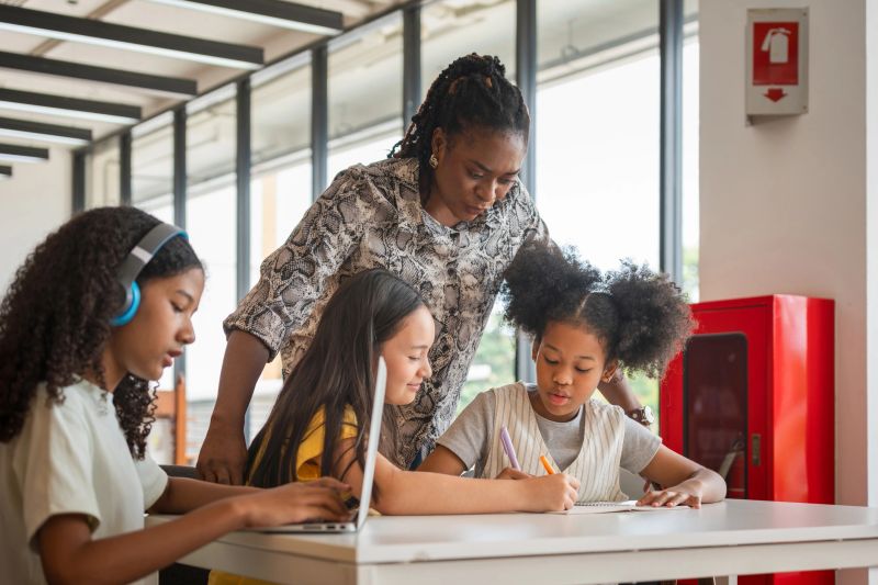 Teacher Guiding Diverse Students in a Modern Classroom, Educator Assisting Children with Schoolwork and Laptops, Multi-ethnic Group of Kids Learning Together with an Instructor