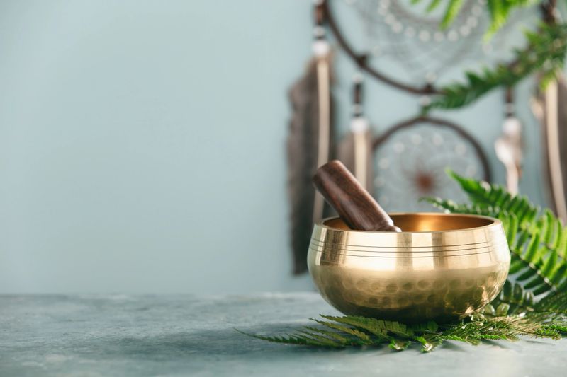 A golden singing bowl with a wooden mallet surrounded by green leaves and spiritual decor. Ideal for wellness, meditation, and holistic healing concepts.