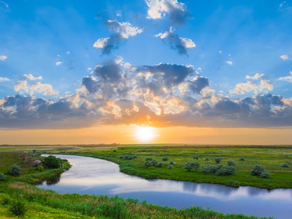 Serene sunset over a winding river and lush green fields with scattered bushes.