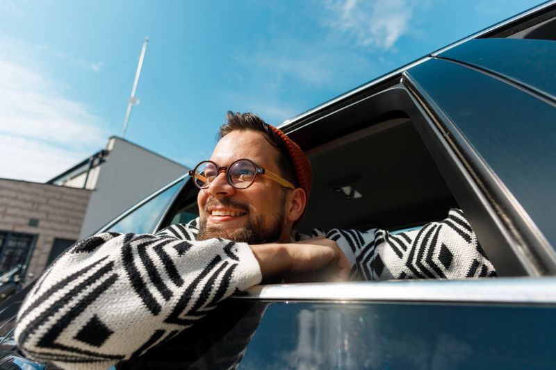 Excited young man cheerfully leaning out of a car window, raising arms in celebration, smiling widely under a sunny sky in an urban setting.
