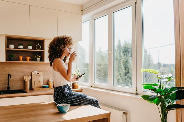 Woman enjoying a drink by the kitchen window with a phone in hand.