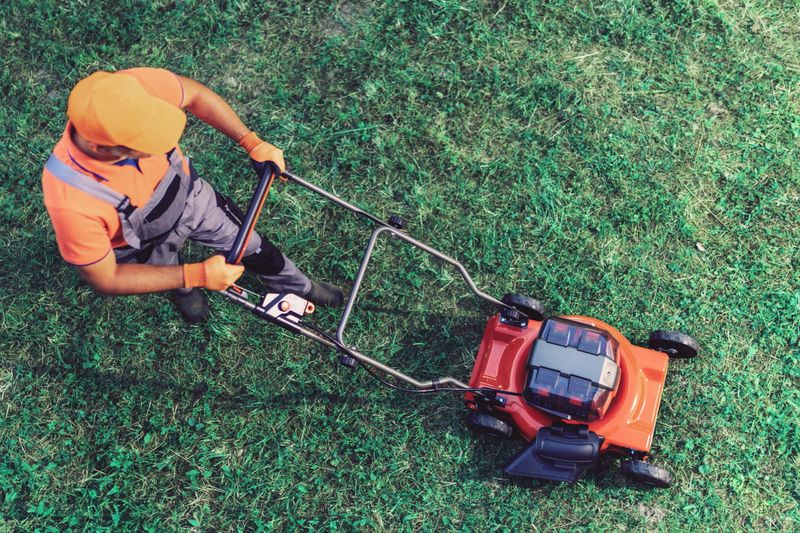 A man is mowing a green lawn with a push mower under clear skies. He wears safety gloves and an orange cap for protection during this outdoor activity.