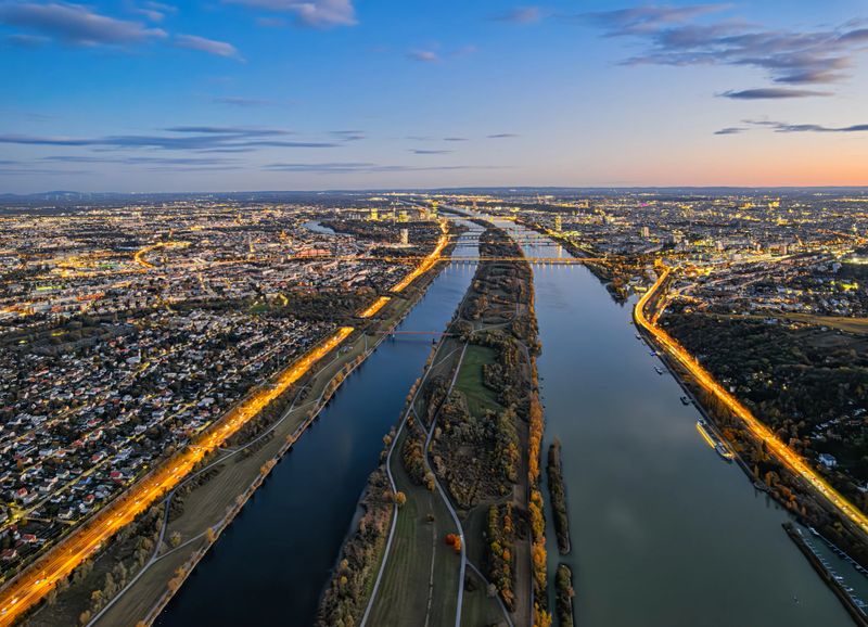 Illuminated streets and cityscape of Vienna stretch along Danube River under colorful evening sky at blue hour.