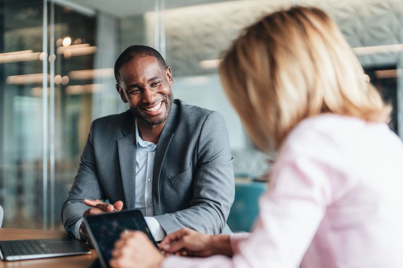 Shot of two confident business persons sitting on a desk in the office and sharing ideas. Businessman and businesswoman in meeting using laptop and discussing business strategy and supply chain.