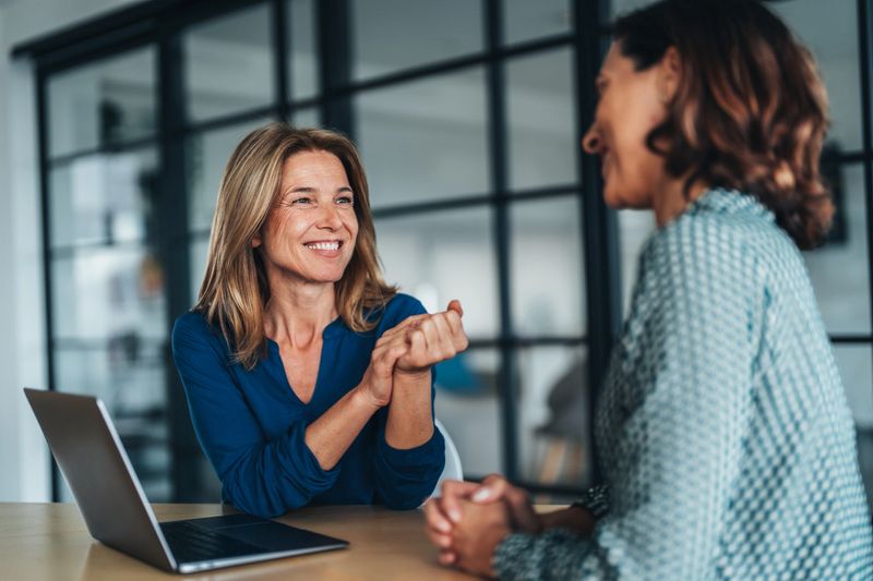 Two women having a focused and friendly conversation at a desk. They wearing formal businesswear, smiling and gesturing with hands, explaining something in a discussion. One woman talking and The other woman, listens attentively while using a laptop. Mentorship, collaboration, or client consultation.