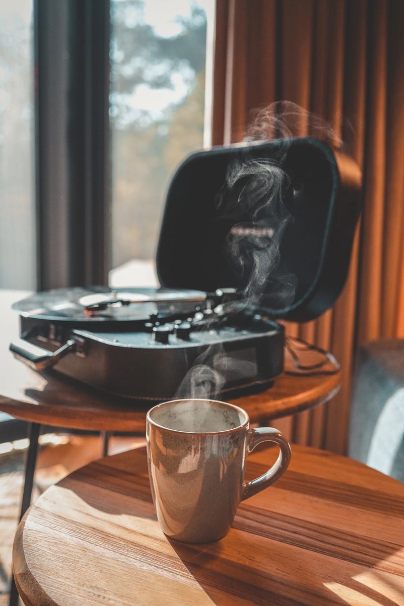 Close-up of a steaming coffee mug on wooden table with a vinyl record player in the background, cozy sunlight streaming into rustic interior.