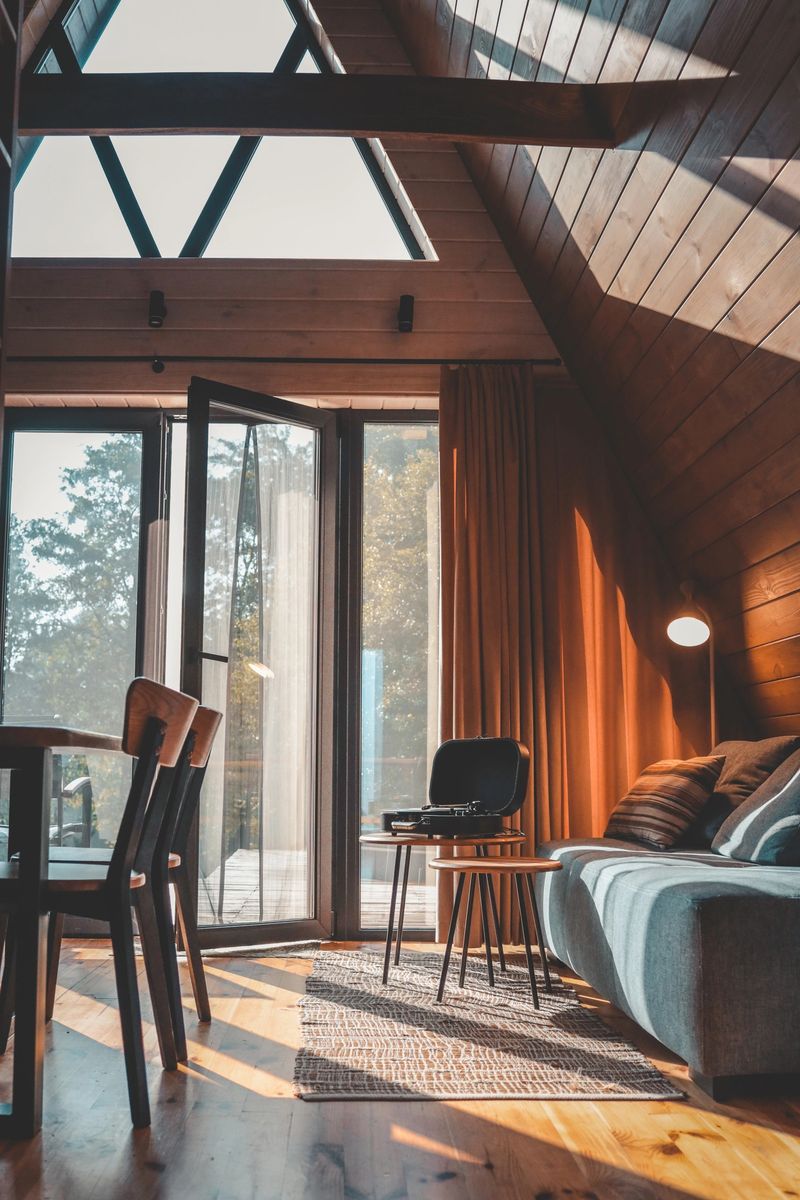 Bright cozy living room in wooden cabin with open door, sunlight on floor, grey sofa, striped pillows, and vintage turntable on small table.