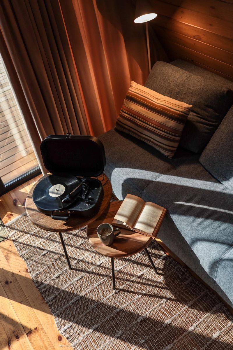 Top-down view of a cozy reading and music corner in a sunlit cabin, featuring an open book, cup of coffee, and a vinyl record player on wooden side tables near a gray sofa.