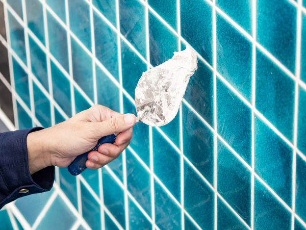 A hand holding a tool near blue tiled wall applying grout or plaster.