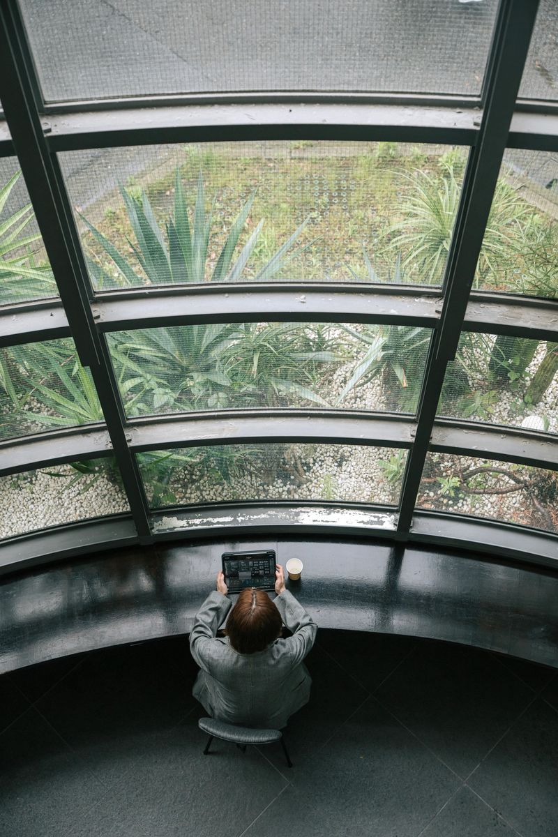 Overhead view of a Japanese businesswoman using a tablet while seated in a curved modern office lounge with large panoramic glass windows overlooking a garden. A coffee cup rests on the bench beside her, creating a calm, productive atmosphere.