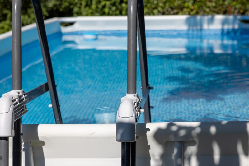 An inviting above-ground swimming pool with clear blue water glistens in the afternoon light, featuring sturdy metal ladder steps for easy access. Surrounded by lush green hedges, this modern pool creates a refreshing oasis perfect for urban or suburban gardens. The scene highlights relaxation, family fun, and summer leisure, ideal for illustrating contemporary outdoor living and home improvement concepts.