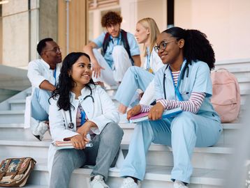 Medical students in scrubs and white coats chatting on steps outdoors.