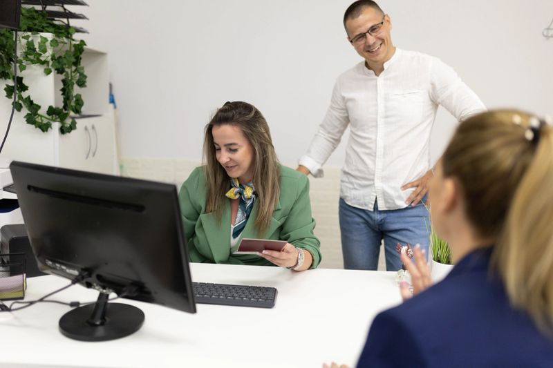 A travel agent and his employee sit at a desk with a client, reviewing the client's passport while the agent demonstrates how to make a reservation on the desktop computer.
