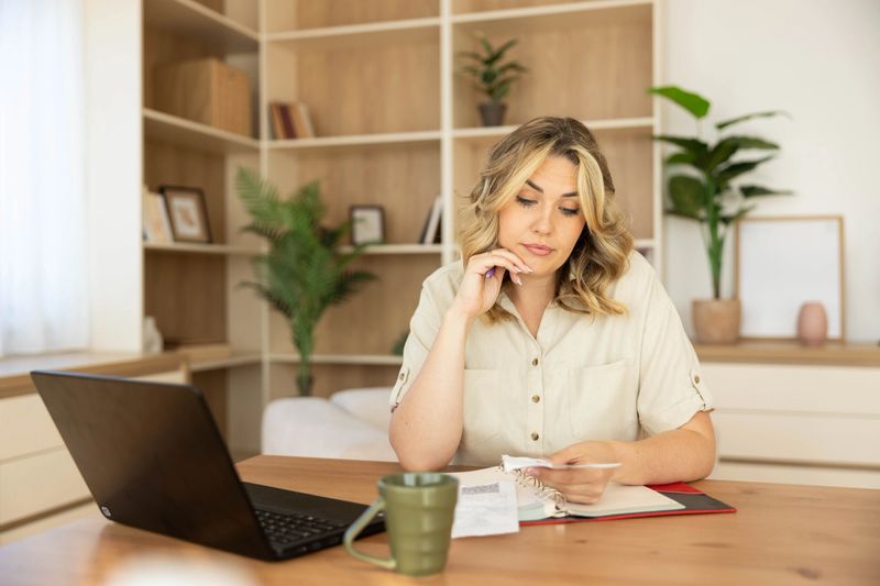 A focused woman sits at a desk surrounded by plants, papers, and a laptop, deep in thought about her work tasks or ideas.