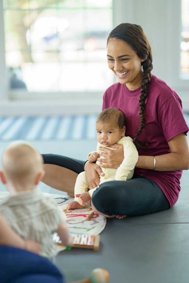 A mother joyfully interacting with her baby and a toddler playing on the floor during a child development bonding session indoors.