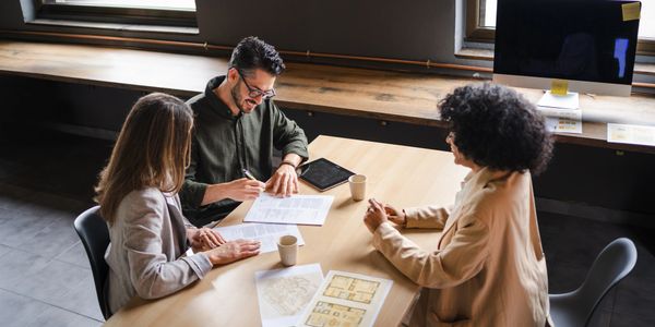 Three professionals discussing documents in a modern office with plans and coffee.