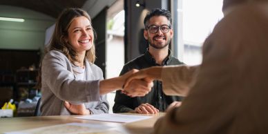 Two people happily shaking hands across a table in a meeting.