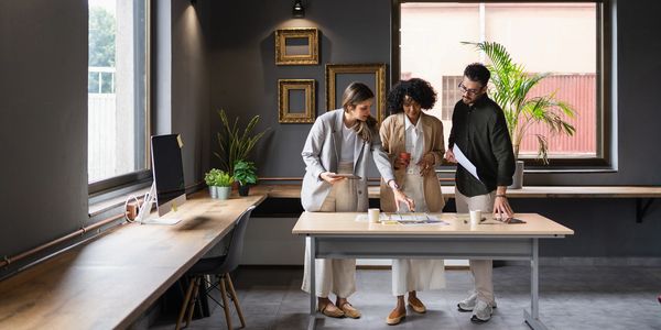 Three colleagues discuss documents in a modern office with dark walls and large windows.