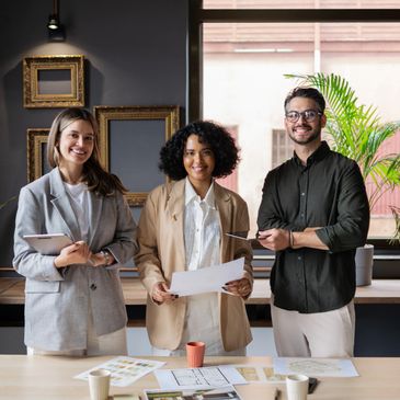 Three professionals smiling and standing behind a desk with documents and tablets.