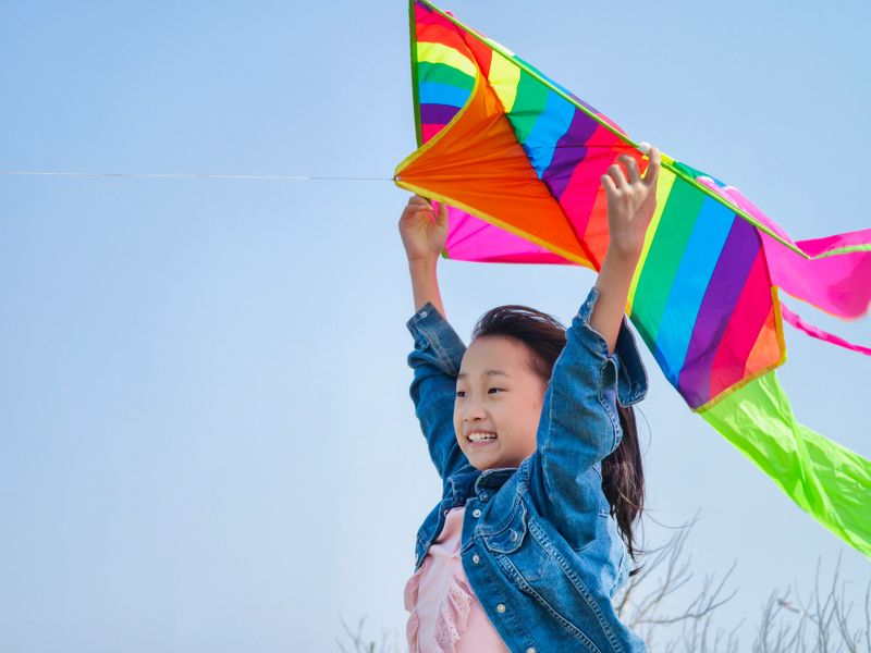 A 9 years old Asian girl flying a kite in the prairie with a background of well-lit sky and grass. Happy childhood life, education. Leisure. Entertainment background.