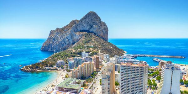 Coastal cityscape with a prominent rocky hill and clear blue sea under a bright sky.