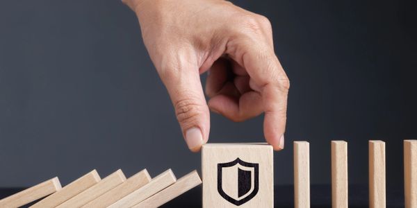 Hand stopping dominoes with a shield symbol block, representing protection.
