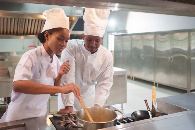 African American Female and Black male chefs in white uniforms and hats stirring pot together, teamwork in modern professional kitchen, culinary training.