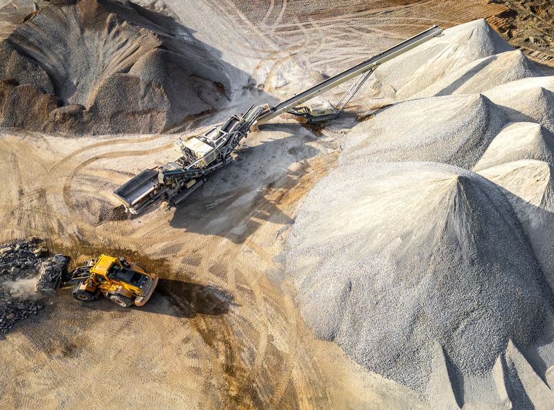 An aerial view of a mining site featuring large gravel piles, a yellow excavator, and industrial machinery under bright daylight with earthy tones.