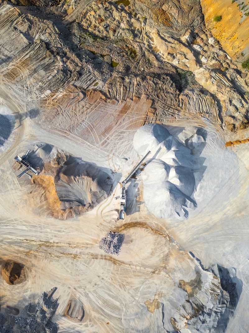 Aerial view of an industrial quarry featuring large gravel piles, heavy machinery, and rugged terrain under bright sunlight and clear skies.