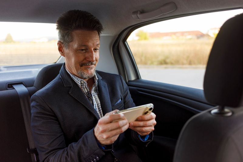 A smiling man is engaged with his smartphone in the backseat of a car, suggesting a pleasant commute. He holds the device horizontally, focused on content that brings him joy, whether it's gaming, watching videos, or browsing the internet. This image captures the modern reality of making the most of travel time through digital entertainment and connectivity.