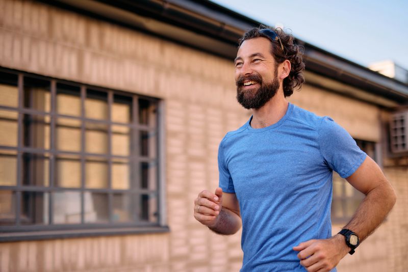 Mid adult man with a beard running energetically in an urban environment, wearing a blue t-shirt, sunglasses, and a wristwatch, smiling while looking away with a sense of joy
