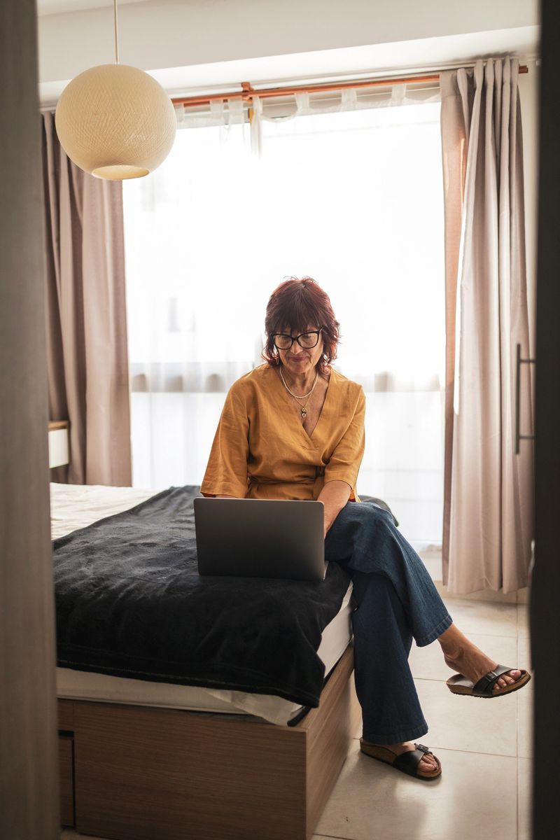 A mature woman sitting on a bed working on a laptop in a bright room, reflecting focus and productivity. Neutral decor and soft lighting create a peaceful and comfortable atmosphere.