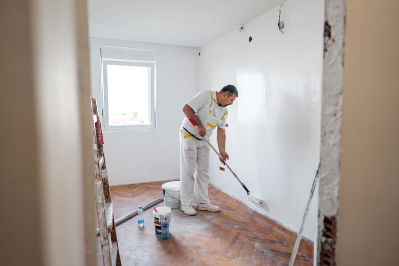 Mature worker painting the walls with a paint roller in an empty apartment under renovation.