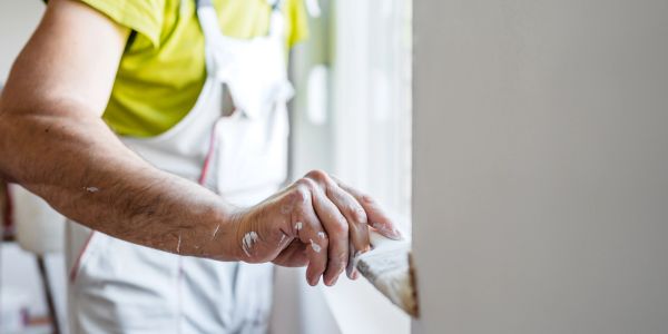 Person painting a window frame with white paint, wearing white overalls and a yellow shirt.
