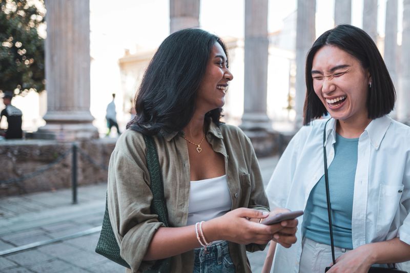 Two female tourists are sharing a funny moment in front of the columns of san lorenzo in milan, italy, using a smartphone