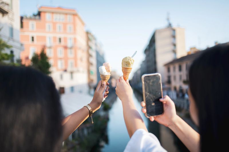 Two tourists enjoying ice cream cones while capturing memorable moments in milan, italy, on a sunny afternoon filled with fun