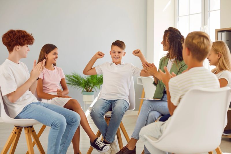 Friendly young woman conducting psychological training for a group of elementary school children sitting in a circle in the classroom applauding and looking to the boy making power winner gesture.
