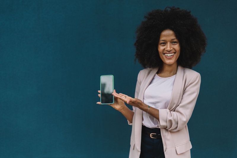 A joyful woman with curly hair is displaying a smartphone with a bright smile, standing against a blue wall, showcasing her confidence and engagement with technology.