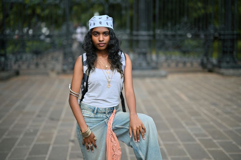 A teenage girl stands confidently on a tiled pavement, striking a powerful pose. She wears street style relaxed clothing. Her confident expression and laid-back style reflect a bold, youthful urban attitude.