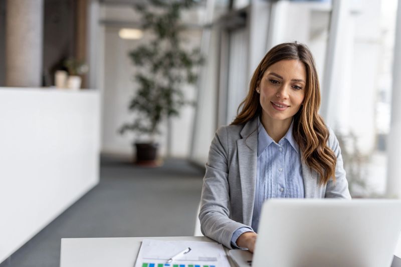 Smiling businesswoman sitting at a modern desk, working on a laptop while reviewing documents and analyzing graphs in a bright and clean office environment