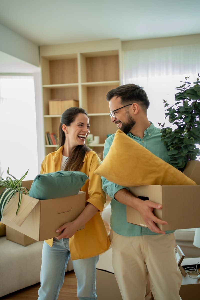 Young couple laughing and carrying boxes filled with cushions and plants while moving into their new home, radiating joy and excitement as they embark on this fresh start together