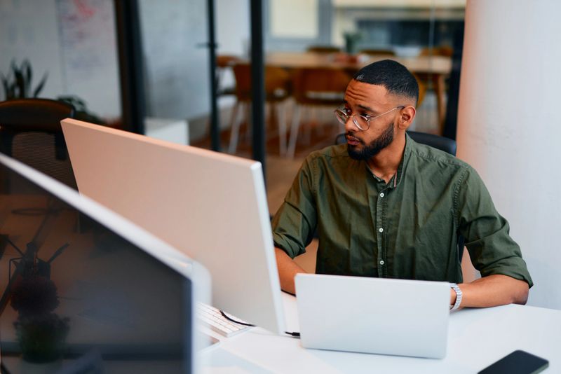 Young adult programmer carefully looking at computer monitor while working on code in modern office, using laptop and desktop computer simultaneously for efficient software development